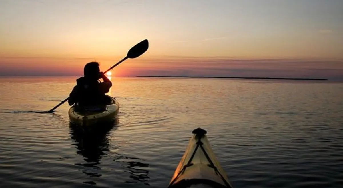 A serene water view at sunset with a single kayaker gliding across calm waters. The sky glows with sunset colors, reflecting on the water and surrounding landscape—perfectly capturing tranquil nature moments and the nearby Currituck Sound.