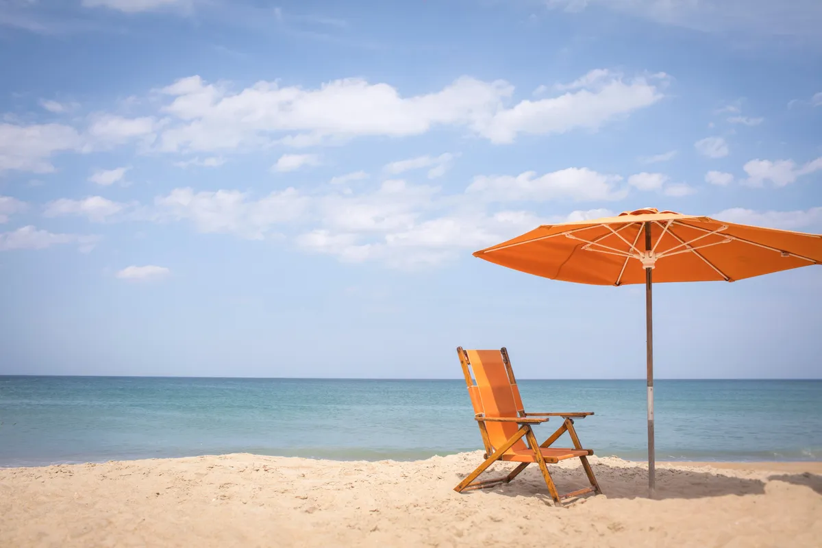 A tranquil stretch of sandy shoreline with a single orange beach chair and matching umbrella facing the calm sea. The scene is set during golden hour, evoking the peaceful ambiance of a quiet morning or sunset at the beach.