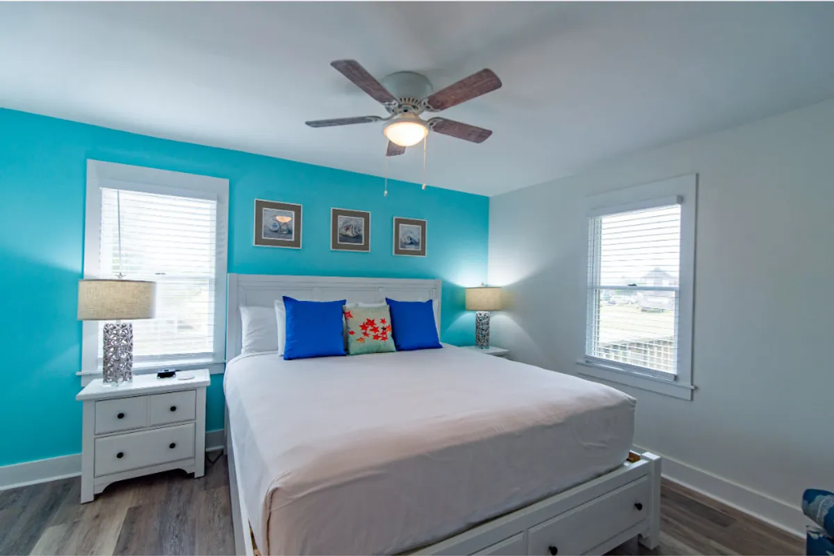 A tranquil bedroom featuring a plush bed adorned with decorative pillows, a calming blue accent wall, framed pictures, and nightstands with softly glowing lamps. The space radiates natural light, creating a restful and inviting atmosphere.