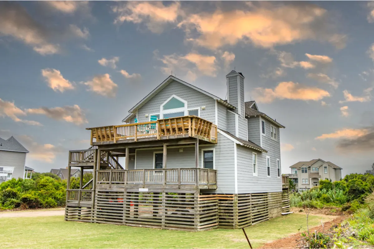 The image showcases the exterior of a large single-family house in Duck, NC, with a spacious deck perfect for gatherings. The home's charming siding and expansive windows look out onto lush greenery, evoking a peaceful and inviting atmosphere. The deck is set up for outdoor relaxation and entertaining, suggesting space to watch sunsets, host barbecues, or enjoy the fresh coastal air.