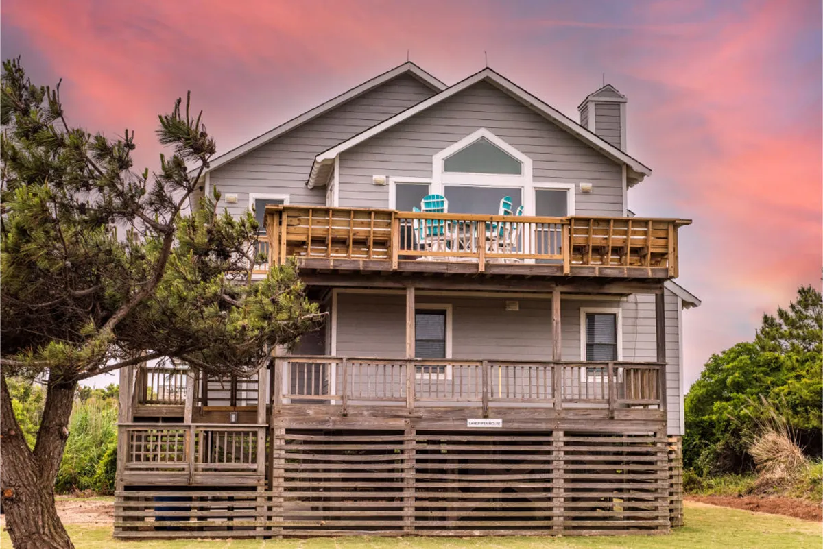 A spacious deck area features comfortable chairs arranged to face the vibrant evening sky, with the house’s façade and lush greenery in the background. Sunlight glows across the wooden boards, inviting visitors to unwind and enjoy the tranquil atmosphere. The seating is ideal for relaxing, socializing, or simply absorbing the sunset views over the Currituck Sound.