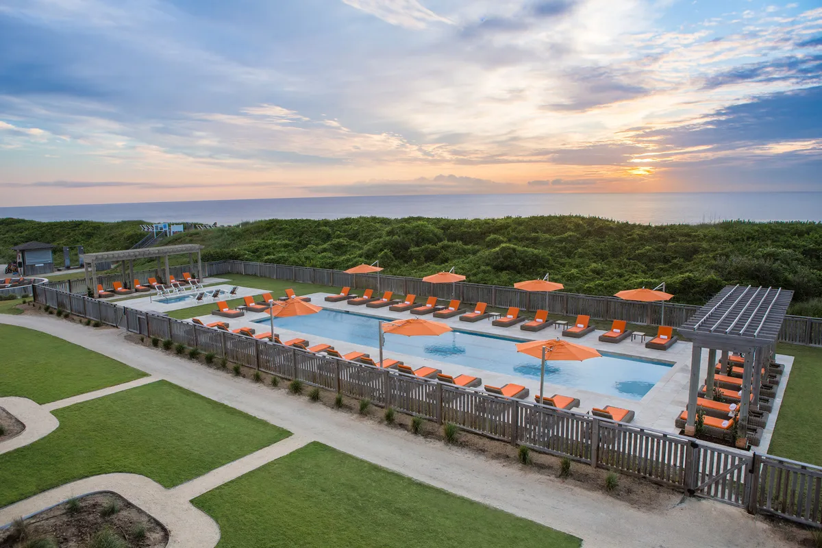 A lively poolside scene at sunset, featuring lounge chairs, orange umbrellas, and people socializing around a shimmering swimming pool. The glowing sunset adds warmth and highlights the resort-style amenities available just steps from the beach.