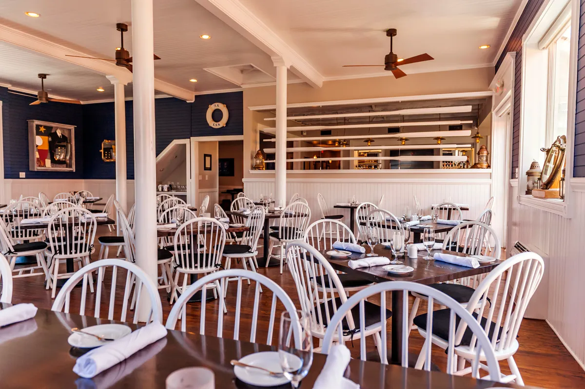 A cheerful dining area with white wooden chairs and dark tables, complemented by blue walls adorned with local artwork. A bar area is visible in the background, inviting social interaction and celebration. The ambiance is lively and relaxed, encouraging group meals and special occasions.