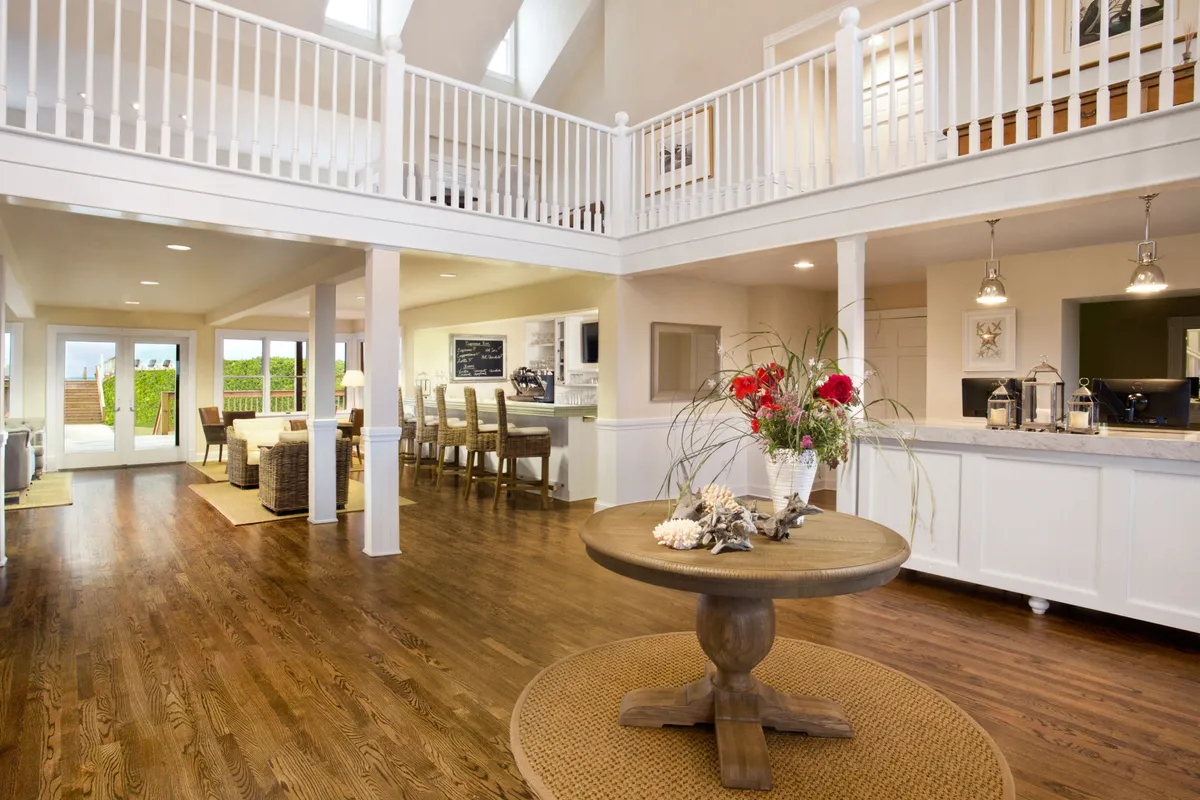 The image shows a circular table with a floral centerpiece, adjacent to an open kitchen area. Large windows provide views of the outdoors, bathing the space in sunlight and reinforcing the open, airy feel. The layout is perfect for casual dining and entertaining.