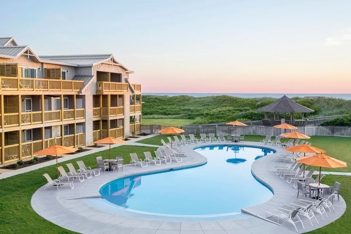 A cheerful scene of the resort’s swimming pool surrounded by orange umbrellas, lush lawns, and balconies overlooking the area. Guests can be seen lounging, swimming, and gathering by the pool, evoking a sense of relaxation and social enjoyment.