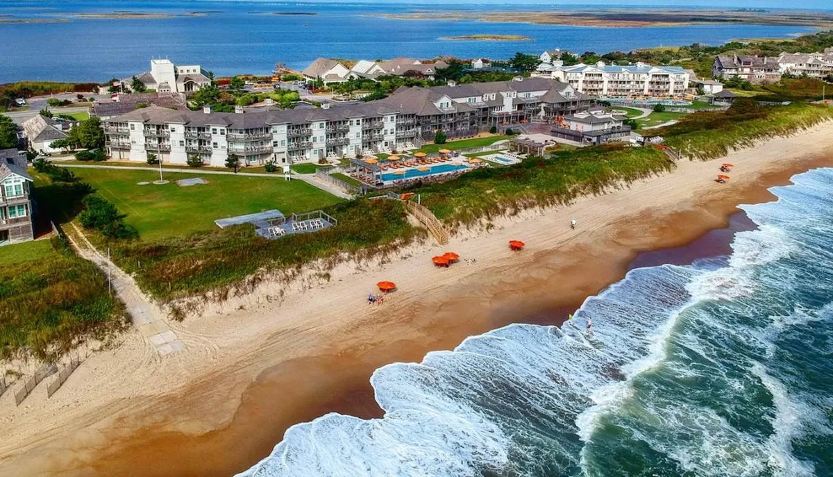 A sweeping exterior shot showcasing the beachfront hotel, a sparkling swimming pool, colorful umbrellas, and waves breaking on the shore. The image captures both the proximity to the beach and the resort's welcoming, resort-style amenities.