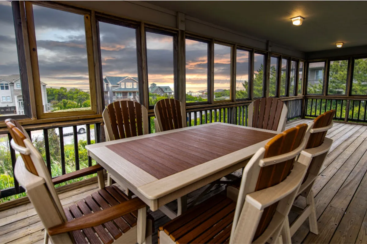 This image features a dining area with wooden chairs and a large table set in front of expansive windows that capture a vibrant sunset. The setting exudes warmth, with natural light filling the room and offering a tranquil view of the outdoors. The scene is set for family gatherings, evening meals, or relaxed game nights, all while surrounded by the beauty of Duck's natural landscape.