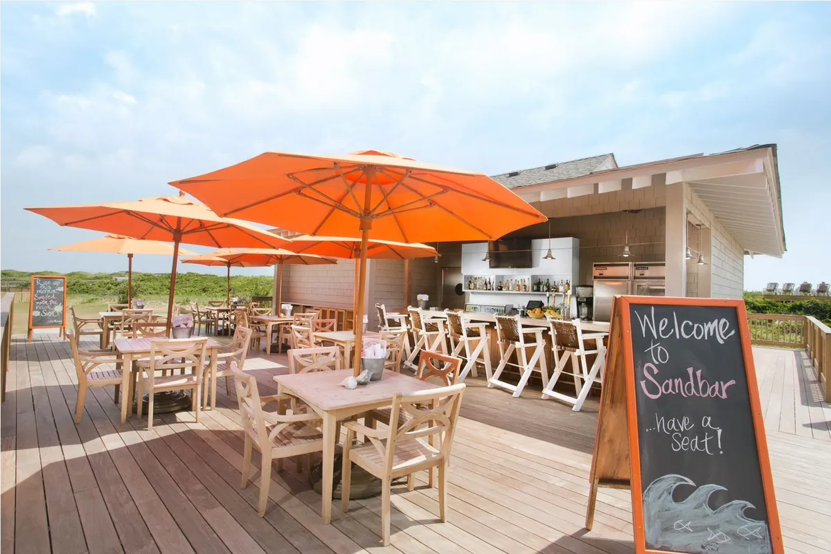 A lively outdoor dining area with bright orange umbrellas, wooden tables, a chalkboard sign, and a casual bar setup. The space feels welcoming and social, perfect for enjoying a meal or drink while staying connected to the outdoors.