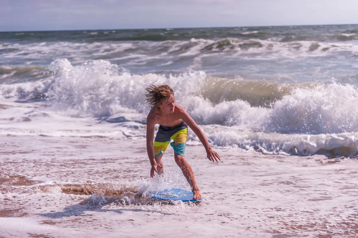 A vibrant exterior scene of a boy riding a boogie board in the surf with waves crashing in the background and sandy beach in the foreground. The playful, sun-soaked atmosphere evokes a sense of active relaxation and connection to the outdoors.