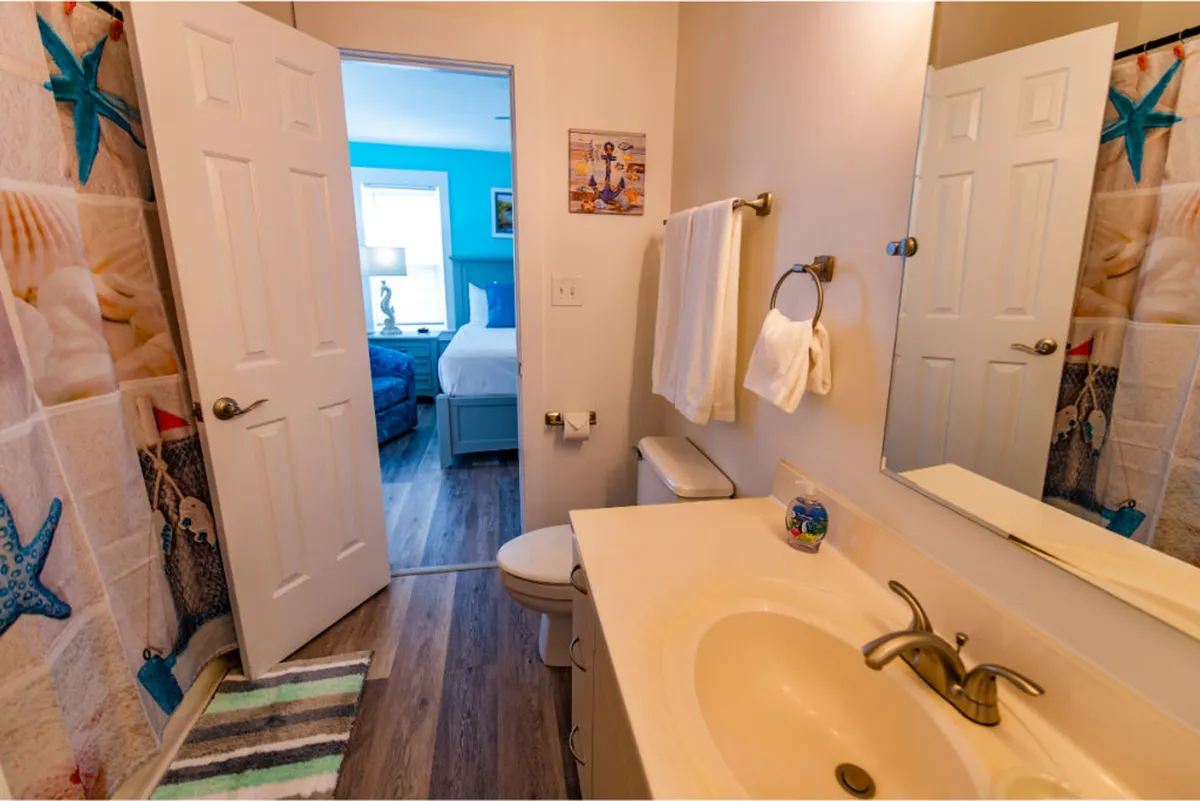 A bright bathroom space with a mirrored vanity, colorful shower curtain, and a view into the adjoining bedroom. The atmosphere is cheerful and calming, suggesting a spa-like bathing experience.