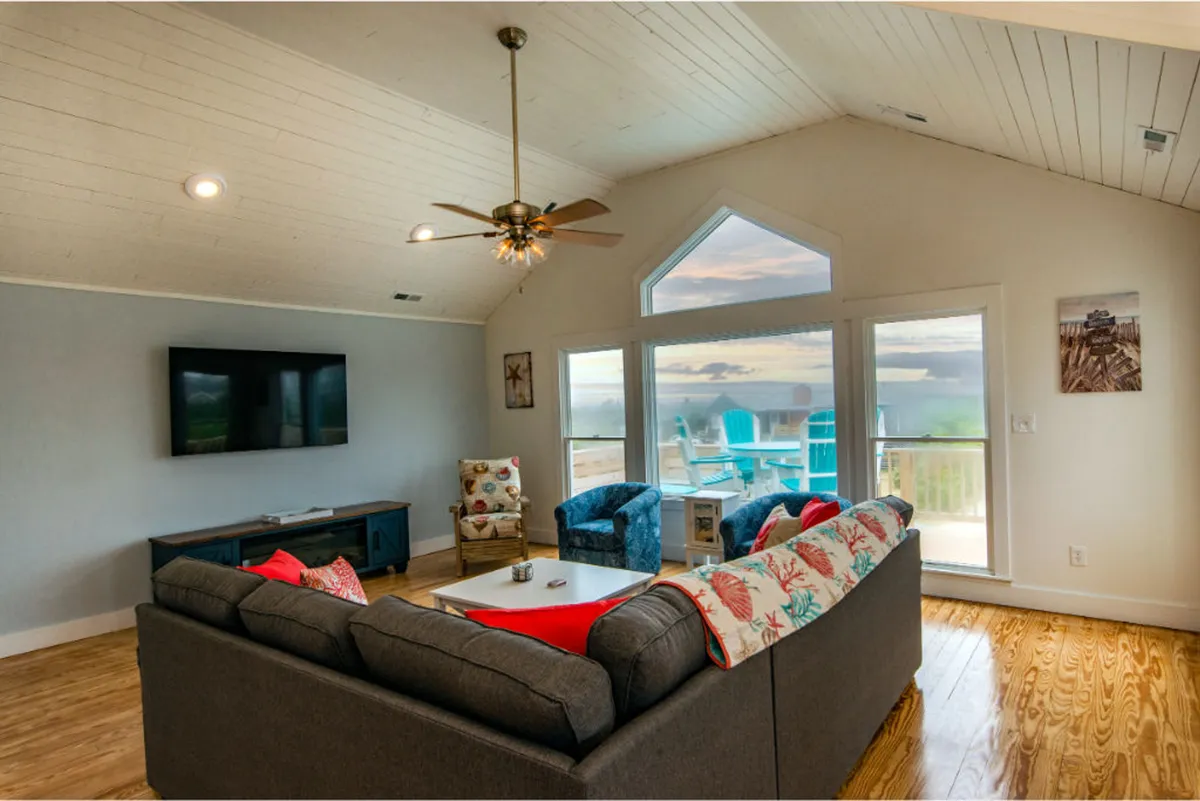 An inviting living room interior featuring expansive windows with a stunning sunset view, a comfortable sectional sofa, and colorful accent chairs. The space is designed for relaxation and taking in the tranquil beauty of the sound.