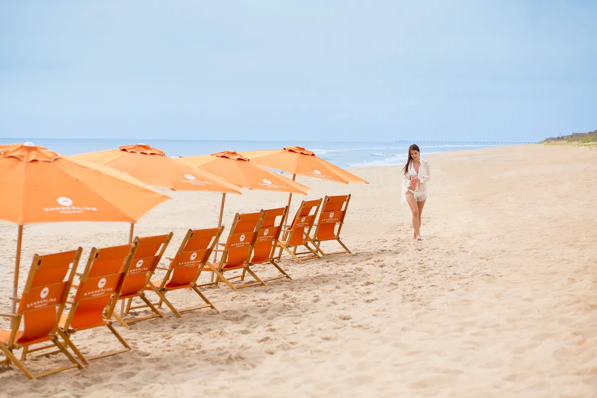 A sun-drenched beach scene with several beach chairs and umbrellas set up near the shoreline. A woman walks along the sand, framed by a wide ocean view, suggesting relaxation, playful activities, and direct access to the water. The lighting hints at golden hour, evoking a sense of sunset or sunrise by the sea.