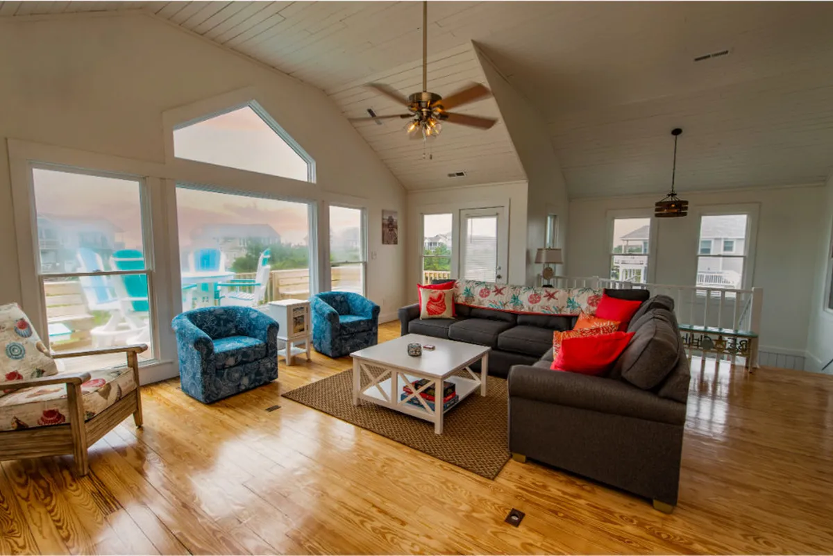 The image features a large sectional sofa with colorful accent chairs arranged around a coffee table, all set in a bright living area with expansive windows offering scenic views. The design encourages relaxation and socializing, with natural light filling the space and a casual, welcoming mood.