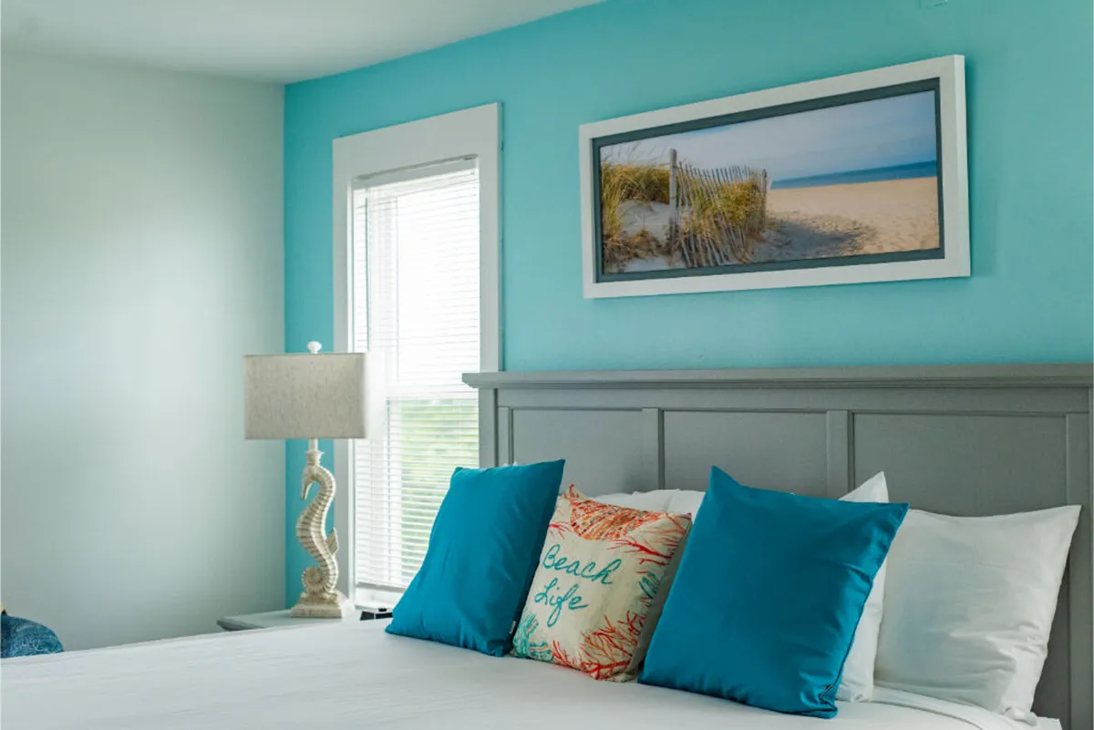 A cheerful bedroom setting with a teal accent wall, decorative pillows, beach-themed artwork, and a bright lamp. Sunlight streams through the windows, offering a cozy nook ideal for both relaxation and waking up refreshed.